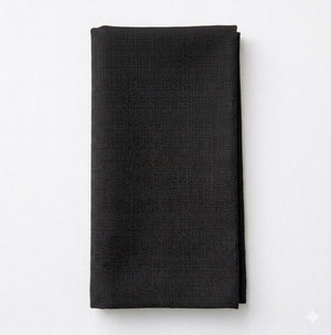 The Amelia Black Linen napkin, crafted with a textured pattern and elegant fabric, highlights quality craftsmanship as it rests neatly folded on a white surface.