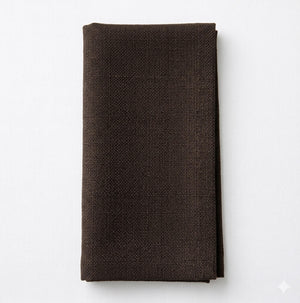 A neatly folded Amelia Dark Chocolate Linen napkin rests on a white surface, providing an elegant, textured fabric accent to any event setting.