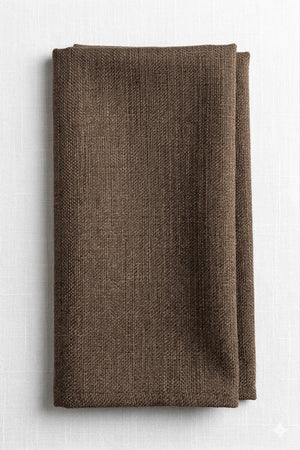 A neatly folded Amelia Light Brown Linen napkin, crafted from elegant fabric, rests on a white, subtly textured surface.