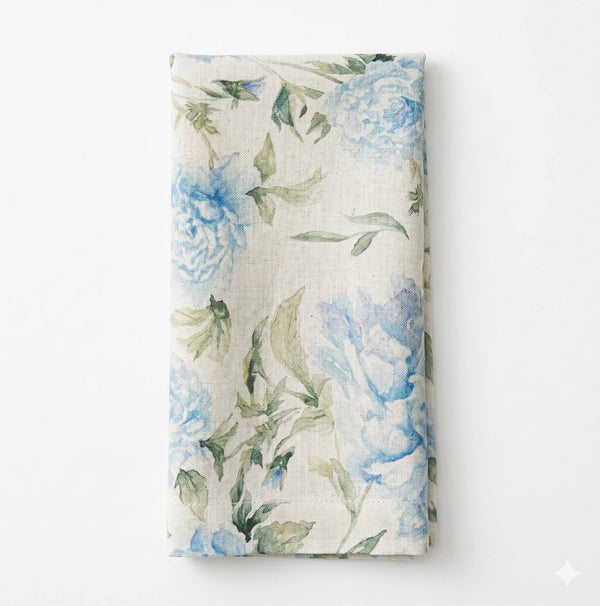The Celeste Linen napkin features a watercolor-style floral pattern with blue flowers and green leaves on white, perfect for elegant table settings.