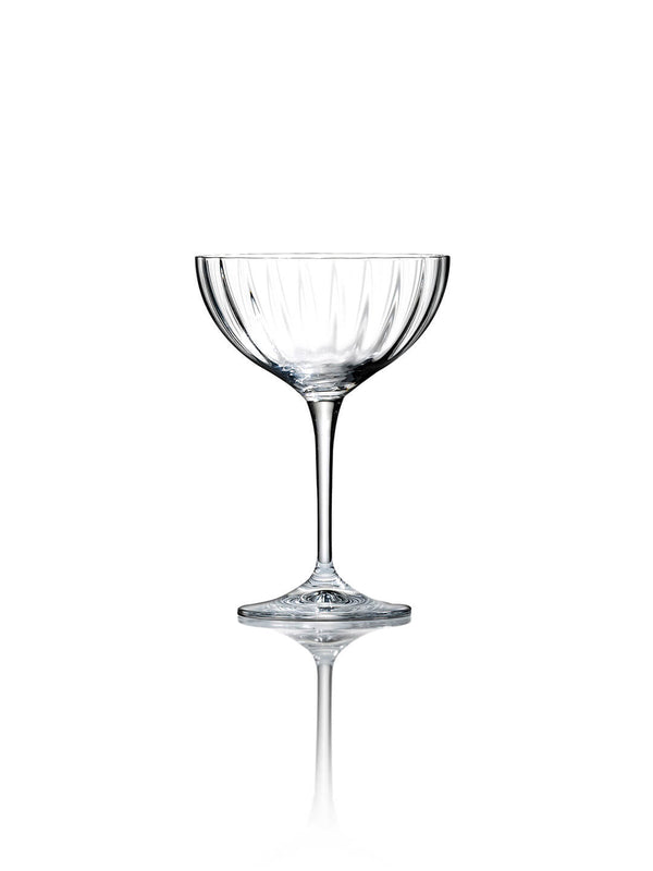 A single empty coupe glass from the Catherine Glassware Range, with a ripple-textured bowl and slim stem, stands on a reflective white surface against a plain white background—perfect for luxury weddings.