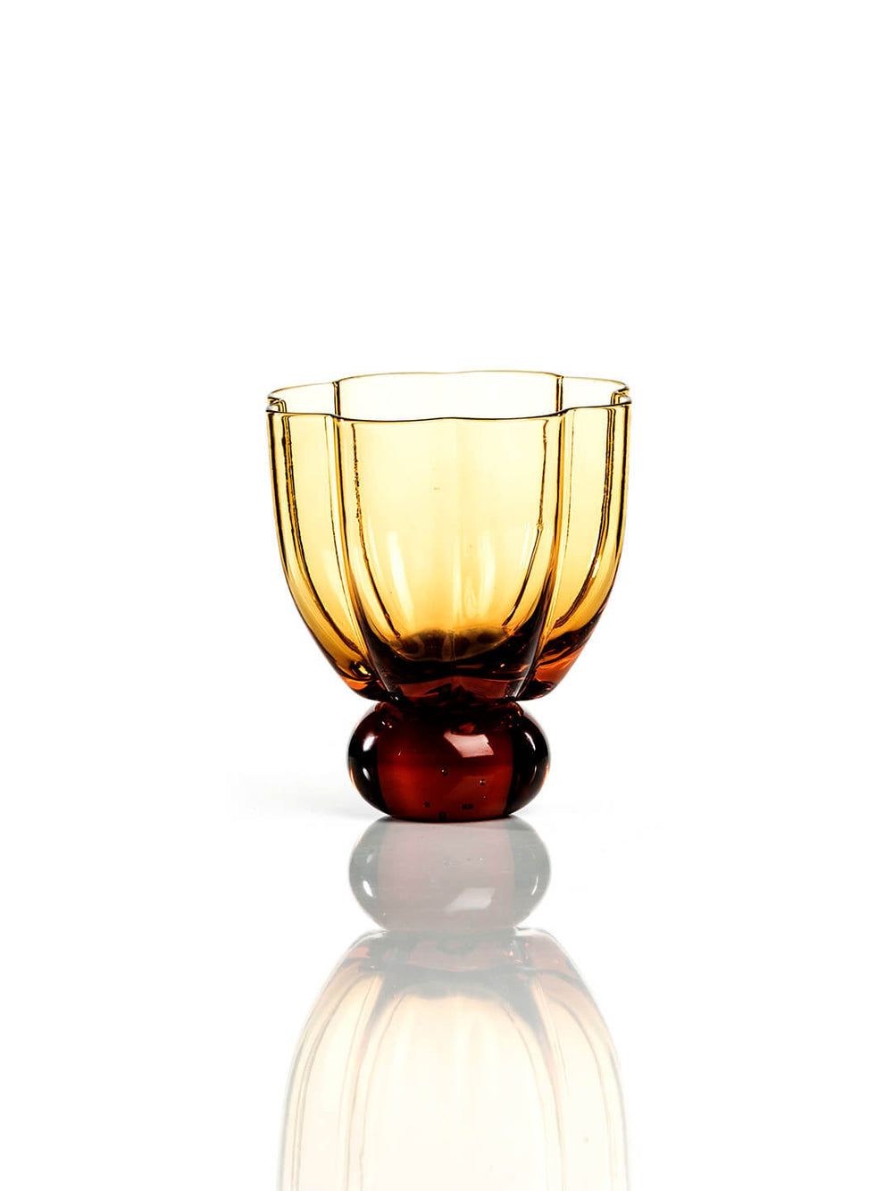 The Flowerbomb Amber Tumbler is a small glass with vertical grooves and a rounded, thick base, designed in an amber color and photographed on a white background with a subtle reflection underneath.