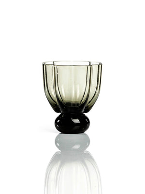 The Flowerbomb Smoke Tumbler is a smoky grey glass cup with a rounded, faceted bowl and thick base, making it striking modern tableware on a reflective white surface.