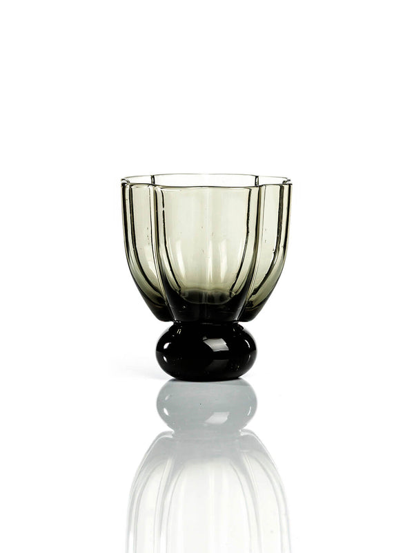 The Flowerbomb Smoke Tumbler is a smoky grey glass cup with a rounded, faceted bowl and thick base, making it striking modern tableware on a reflective white surface.