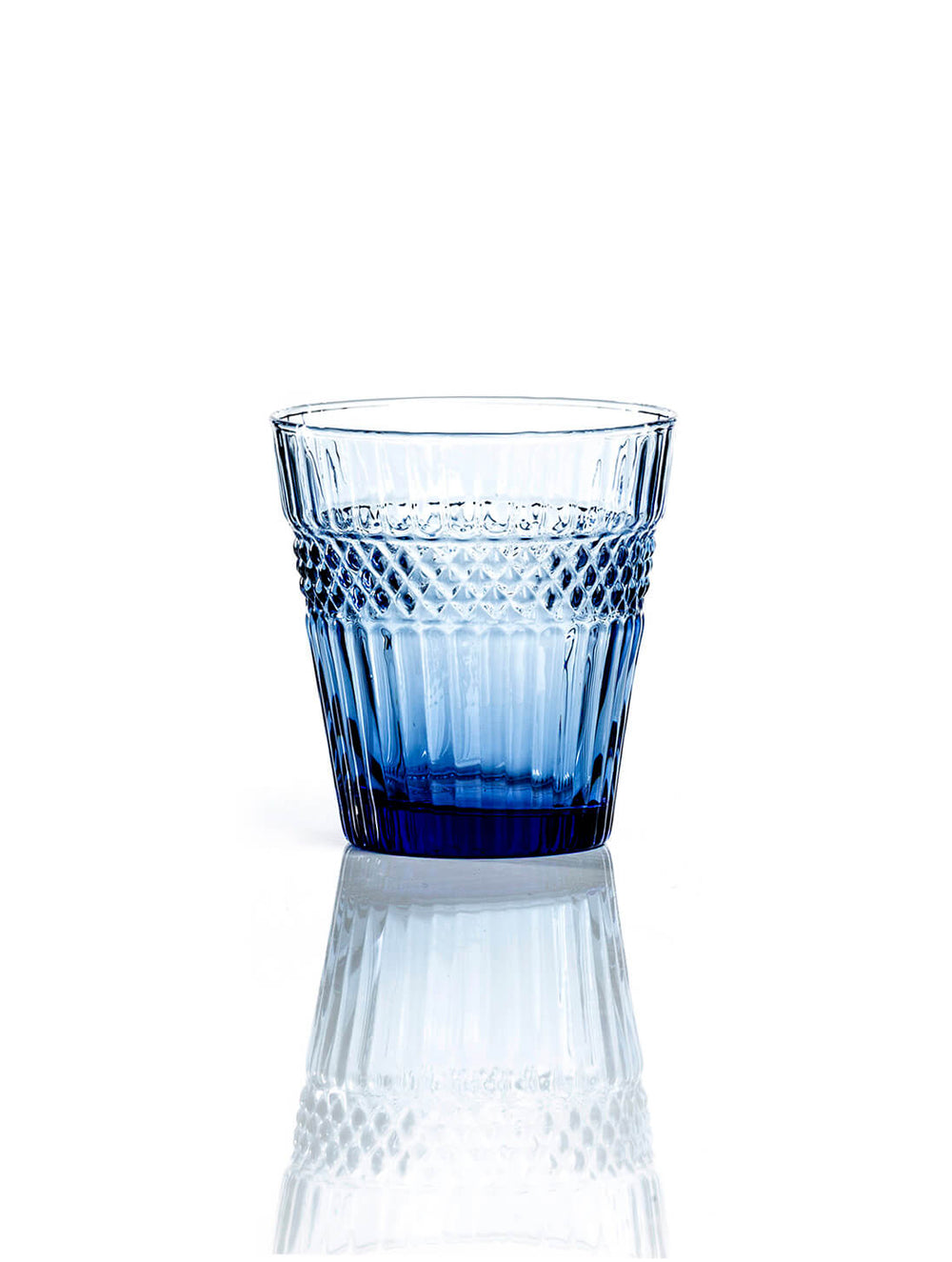 The Margot Blue Tumbler showcases a textured blue-tinted glass and gently flared shape, adding elegance to modern dinner parties. Its unique design stands out against a white background.