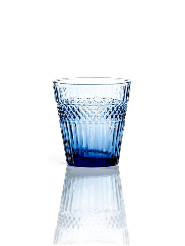 The Margot Blue Tumbler showcases a textured blue-tinted glass and gently flared shape, adding elegance to modern dinner parties. Its unique design stands out against a white background.