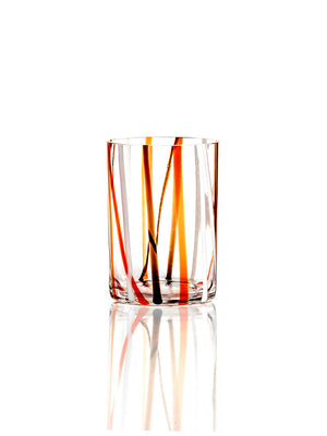 The Reed Orange Tumbler is a clear glass with vertical orange, yellow, black, and red streaks inspired by classic retro glassware, shown on a reflective white surface against a white background.