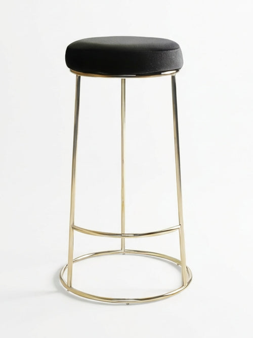 The Astor Bar Stool features a modern design with a round black velvet cushioned seat and a shiny gold metal frame, supported by four slender legs with circular and horizontal connectors.