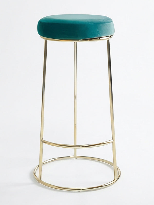 The Astor Bar Stool features a round teal velvet cushioned seat, slender gold metal legs, and a circular footrest. It is set against a plain white background.