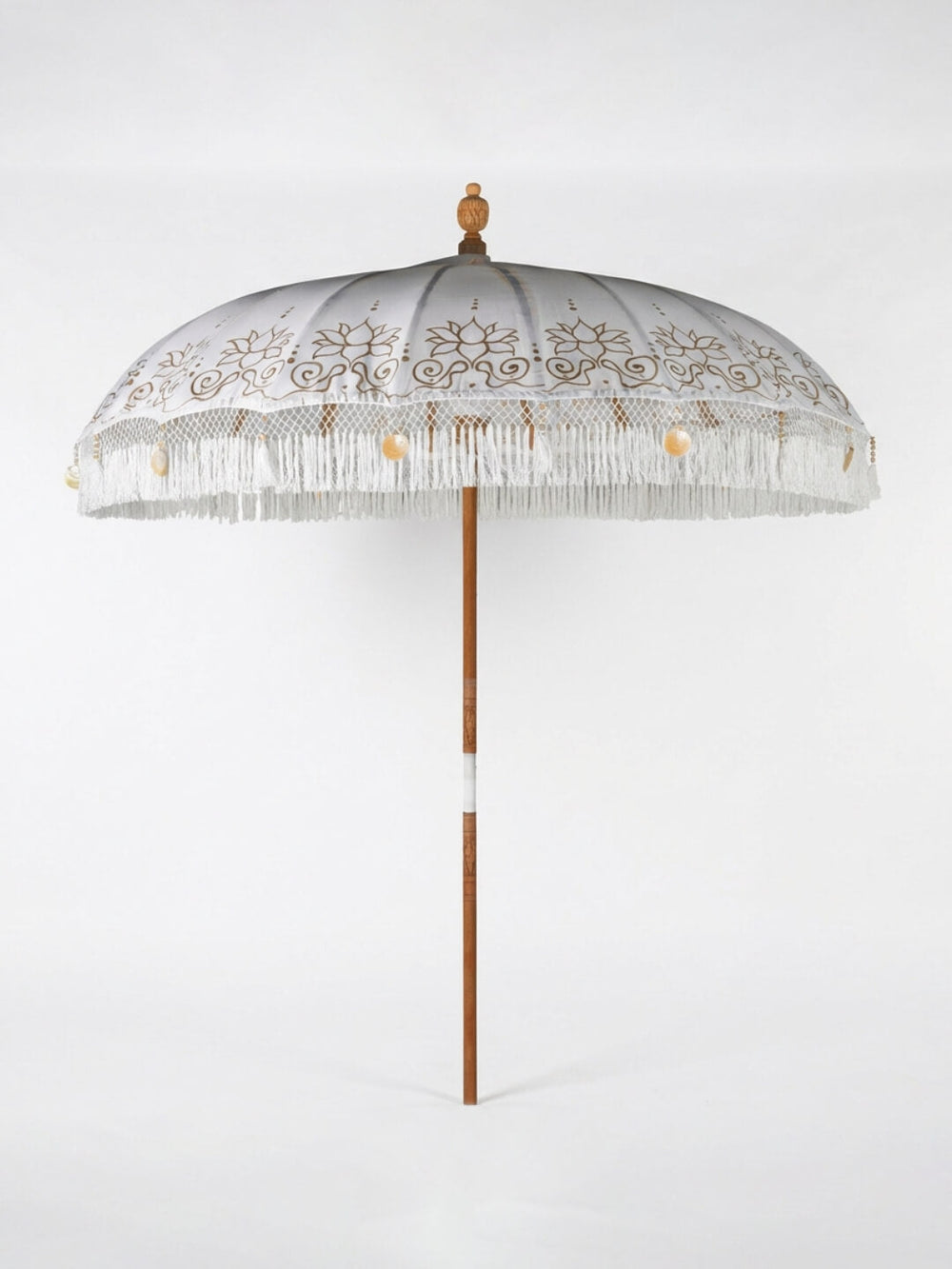 The Bali Dream Parasol features a decorative white handpainted canopy with gold patterns, white fringe, small hanging tassels, and a wooden pole—ideal as bohemian wedding decor or for outdoor events.