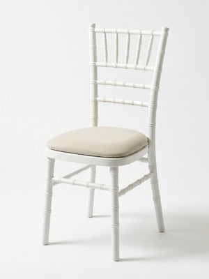 The Chiavari Chair - White features a spindle-style backrest and legs with a light beige cushioned seat, perfect for events. Custom seat pads are available for extra comfort. Shown against a plain white background.