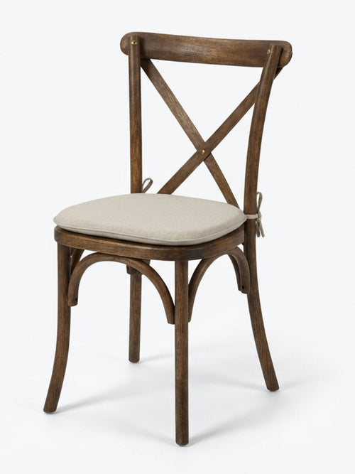 The Cross Back Chair has a wooden frame with a dark brown finish, an X-shaped backrest, and a light beige cushioned seat. Its curved legs and classic design make it perfect for rustic events or wedding chair hire.