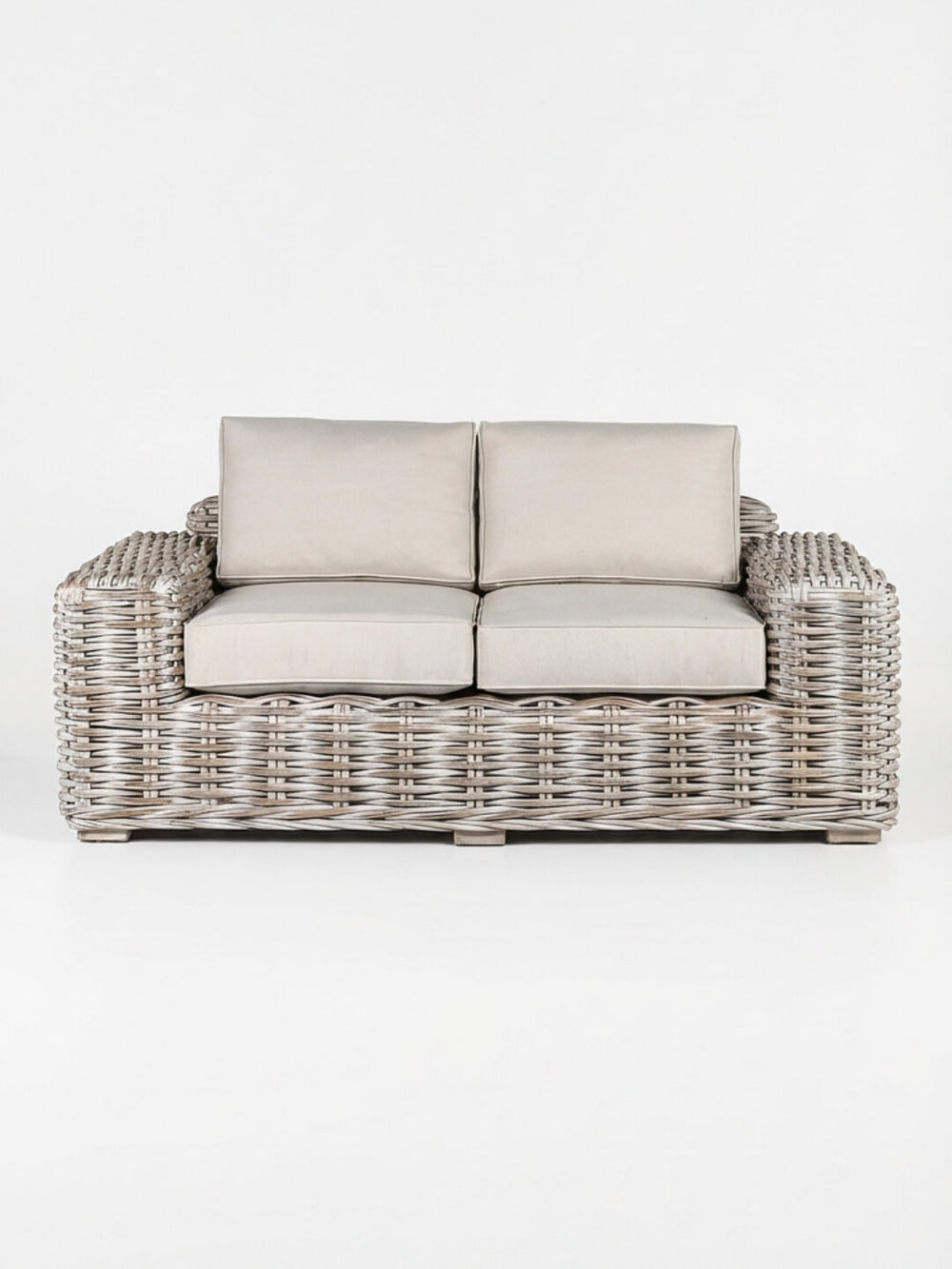 The Driftwood Sofa is a two-seater with a light gray woven rattan frame and plush beige cushions, showcasing coastal charm against a plain white background.