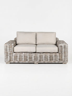 The Driftwood Sofa is a two-seater with a light gray woven rattan frame and plush beige cushions, showcasing coastal charm against a plain white background.