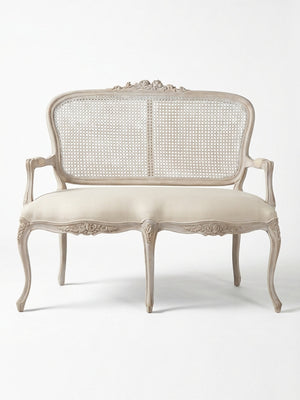 The Fleur Sofa features an antique-inspired carved wooden frame, curved legs and arms, a woven cane backrest, and a beige upholstered seat, highlighting French elegance against a plain white background.
