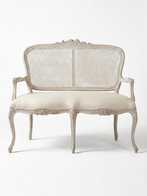The Fleur Sofa features an antique-inspired carved wooden frame, curved legs and arms, a woven cane backrest, and a beige upholstered seat, highlighting French elegance against a plain white background.