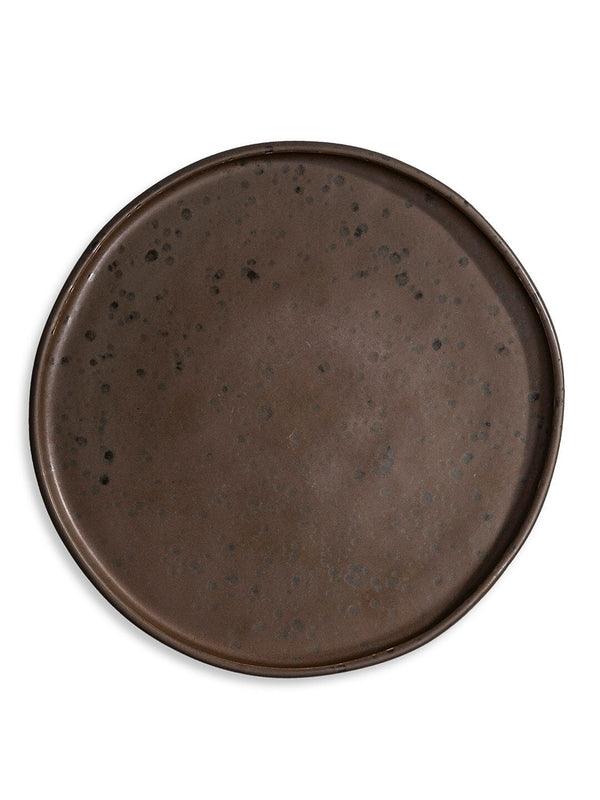The Juniper Charger Plate is a handcrafted, round, dark brown charger with an uneven rim and subtle black speckles that adds rustic charm to any table setting, presented against a white background.