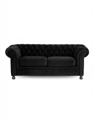 The Luxe Noir Sofa is a black velvet Chesterfield with a tufted backrest, rolled arms, and two seat cushions, shown on a plain white background—an elegant choice for event lounge furniture.