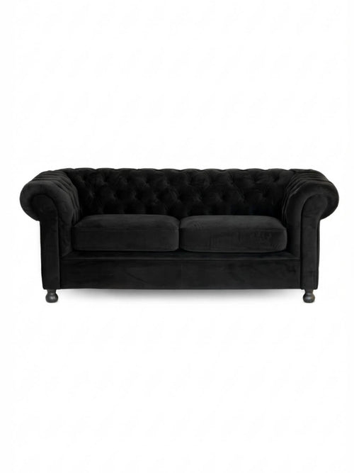 The Luxe Noir Sofa is a black velvet Chesterfield with a tufted backrest, rolled arms, and two seat cushions, shown on a plain white background—an elegant choice for event lounge furniture.