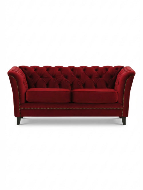 The Merlot Sofa is a luxurious red velvet piece featuring a tufted backrest, flared arms, gold nailhead trim, and four dark wooden legs for a vintage glamour look.