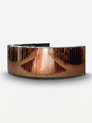 The Mirror Bar features a sleek, curved design with a reflective metallic surface that showcases the surrounding room, from clothing racks to polished wood floors. Set against a plain white background, it’s an ideal customizable event bar for stylish gatherings.