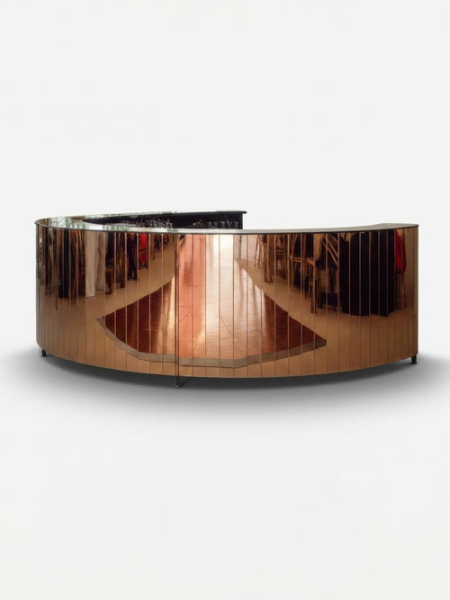 The Mirror Bar features a sleek, curved design with a reflective metallic surface that showcases the surrounding room, from clothing racks to polished wood floors. Set against a plain white background, it’s an ideal customizable event bar for stylish gatherings.