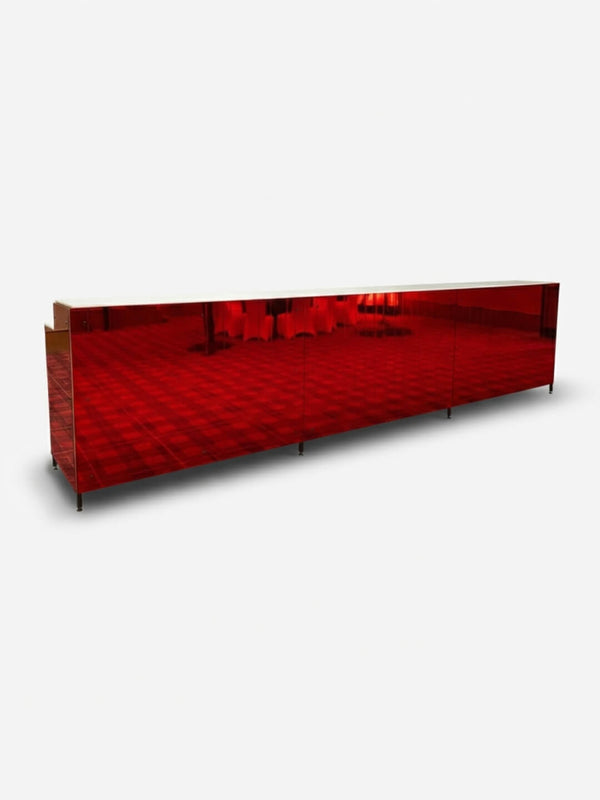 The Mirror Bar, a long rectangular piece with glossy red reflective surfaces, stands on a plain white background, partially reflecting its surroundings.