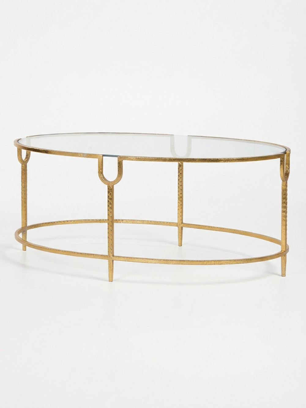 The Monaco Coffee Table showcases an oval glass top on a gold metal base with a textured finish, four legs joined by a circular frame, all set against a plain white background.