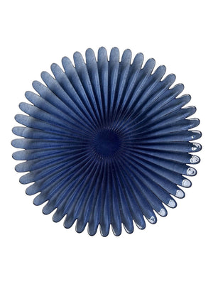 The Petra Blue Charger Plate is round and dark blue with petal-like ridges and a textured center, resembling a stylized flower or fan, displayed on a white background.