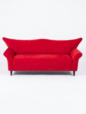 The Rouge Sofa features vibrant red velvet, wide angled armrests, and sleek black legs—its modern design makes a striking statement against any plain white background.