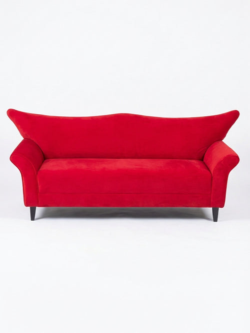The Rouge Sofa features vibrant red velvet, wide angled armrests, and sleek black legs—its modern design makes a striking statement against any plain white background.