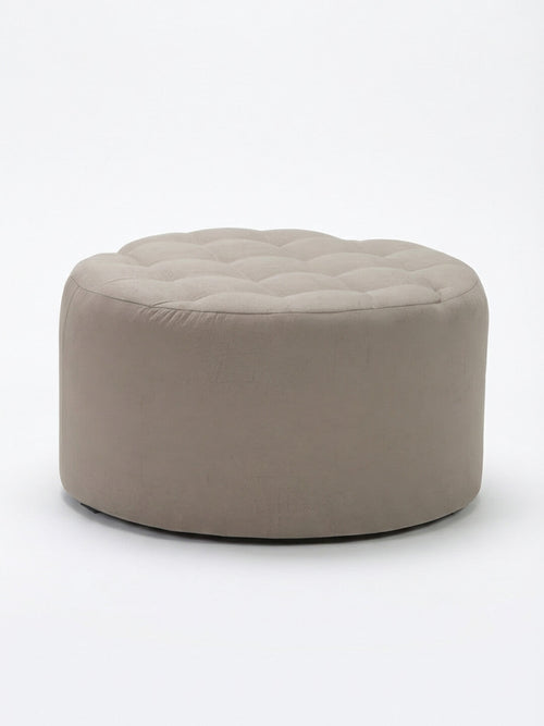 The Sandstone Pouffe is a circular pouffe in light beige linen with a tufted top, offering a touch of elegance and comfort to any space, shown here on a plain white background.