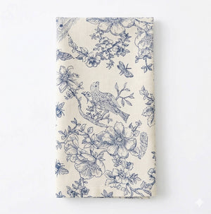 The Chatsworth Linen napkin, in a folded cream hue, displays elegant blue floral and bird motifs for a refined, nature-inspired table setting.
