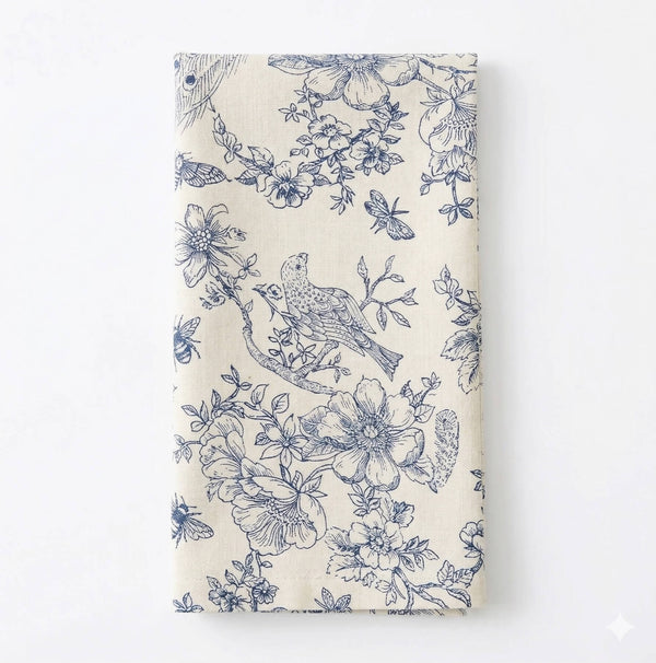 The Chatsworth Linen napkin, in a folded cream hue, displays elegant blue floral and bird motifs for a refined, nature-inspired table setting.