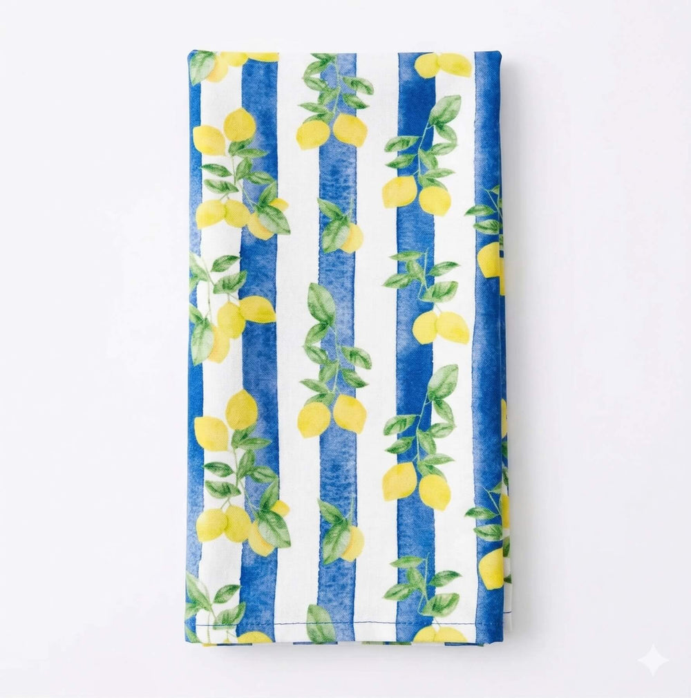 A table draped in Chloe Linen, a white cloth with blue vertical stripes and yellow lemons with green leaves along the stripes, brings vibrant Mediterranean charm to your decor.