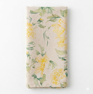 A folded beige Delilah Linen napkin features a watercolor pattern of yellow flowers and green leaves on a plain white background—part of our exclusive collection.