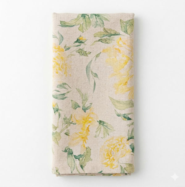 A folded beige Delilah Linen napkin features a watercolor pattern of yellow flowers and green leaves on a plain white background—part of our exclusive collection.