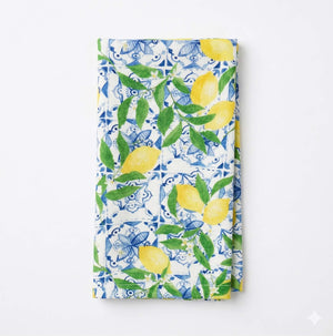The Elena Linen napkin, folded and shown on a plain white surface, features vibrant yellow lemons, green leaves, and blue-and-white decorative tile patterns on a white background.