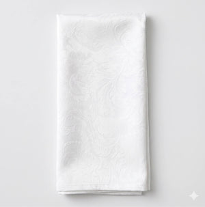 The Evie Linen napkin, neatly folded, features a subtle and elegant white floral damask pattern and is displayed on a plain light background.