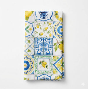 The Giulia Linen napkin showcases Mediterranean elegance with vibrant blue, yellow, and green patterns—lemons and ornate motifs—on a crisp white background.