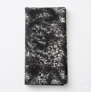 The Isabella Steel Linen, a folded black cloth featuring a textured abstract white and gray pattern, sits on a plain white surface—a striking piece of contemporary table linen.