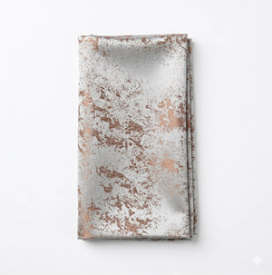A folded napkin from the Isabella Taupe Linen collection features a metallic marbled pattern in copper and silver on an elegant white tablecloth.