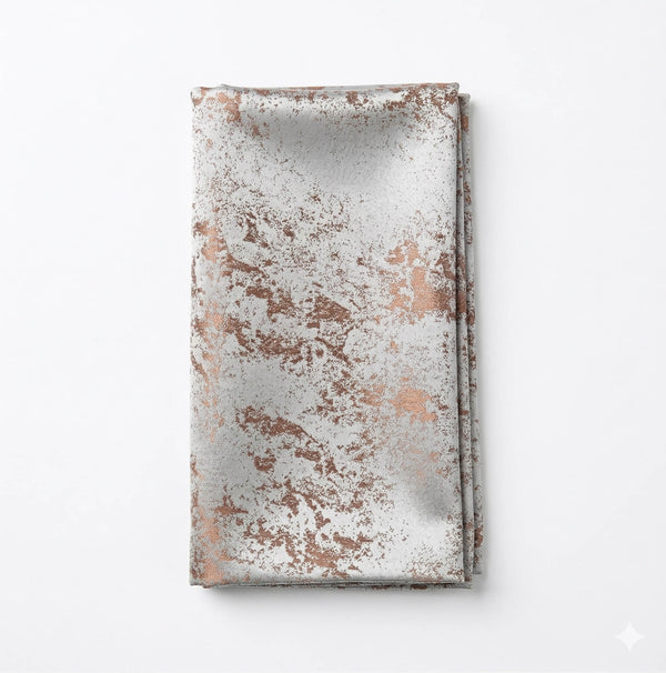 A folded napkin from the Isabella Taupe Linen collection features a metallic marbled pattern in copper and silver on an elegant white tablecloth.