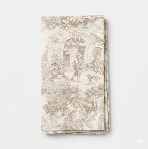 The Jolie Marron Linen is a cream-colored fabric napkin with intricate beige toile patterns in light brown showing people, a house, and nature scenes. It is neatly folded and displayed on a plain white background.