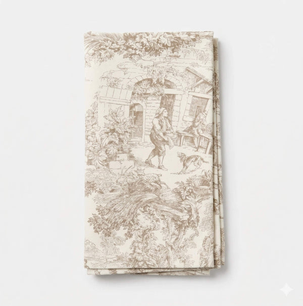 The Jolie Marron Linen is a cream-colored fabric napkin with intricate beige toile patterns in light brown showing people, a house, and nature scenes. It is neatly folded and displayed on a plain white background.