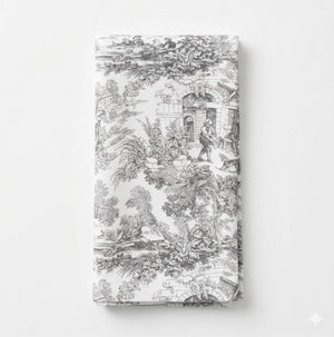 The Jolie Noir Linen features a folded white napkin adorned with intricate black toile patterns of people, trees, and buildings in a vintage style—part of the exclusive collection.