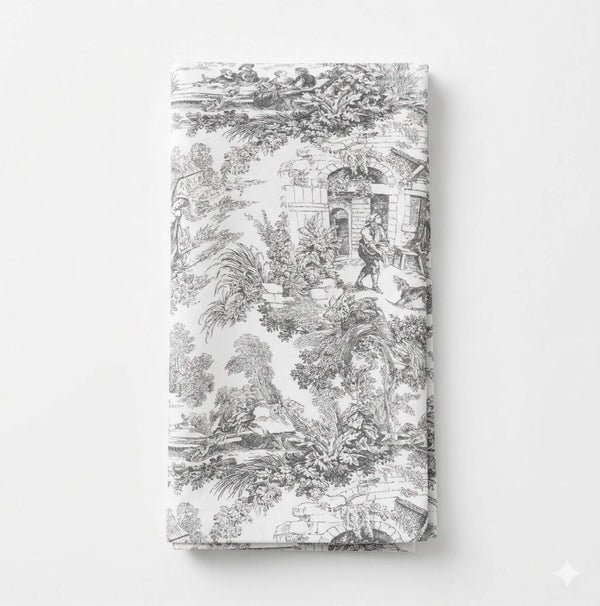 The Jolie Noir Linen features a folded white napkin adorned with intricate black toile patterns of people, trees, and buildings in a vintage style—part of the exclusive collection.