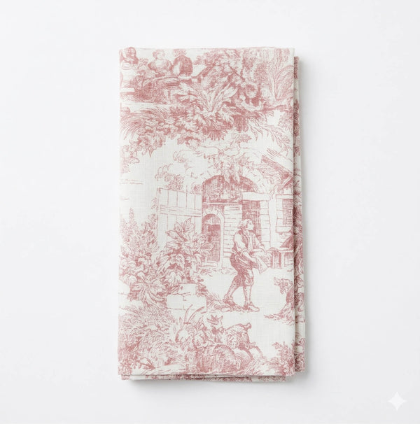 A folded cloth napkin from Jolie Rose Linen, featuring a blush pink toile pattern with pastoral scenes of people and animals—adds romantic charm to any table, ideal for fans of the Jolie Rose Linen collection.