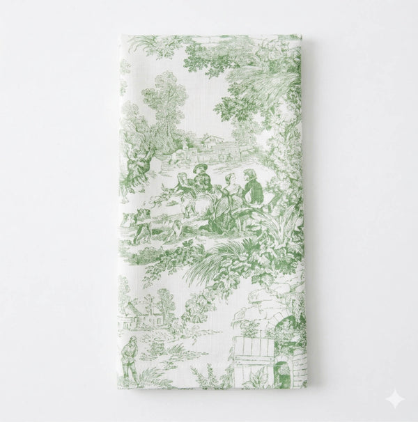 The Jolie Vert Linen napkin showcases a green toile pattern with pastoral scenes—people, trees, and buildings—on a crisp white background, offering timeless sophistication.