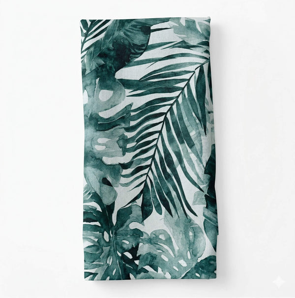 Layla Linen is a folded napkin with large, overlapping tropical leaves in varying green shades on a white background—perfect for adding a fresh touch to your table or event decor.